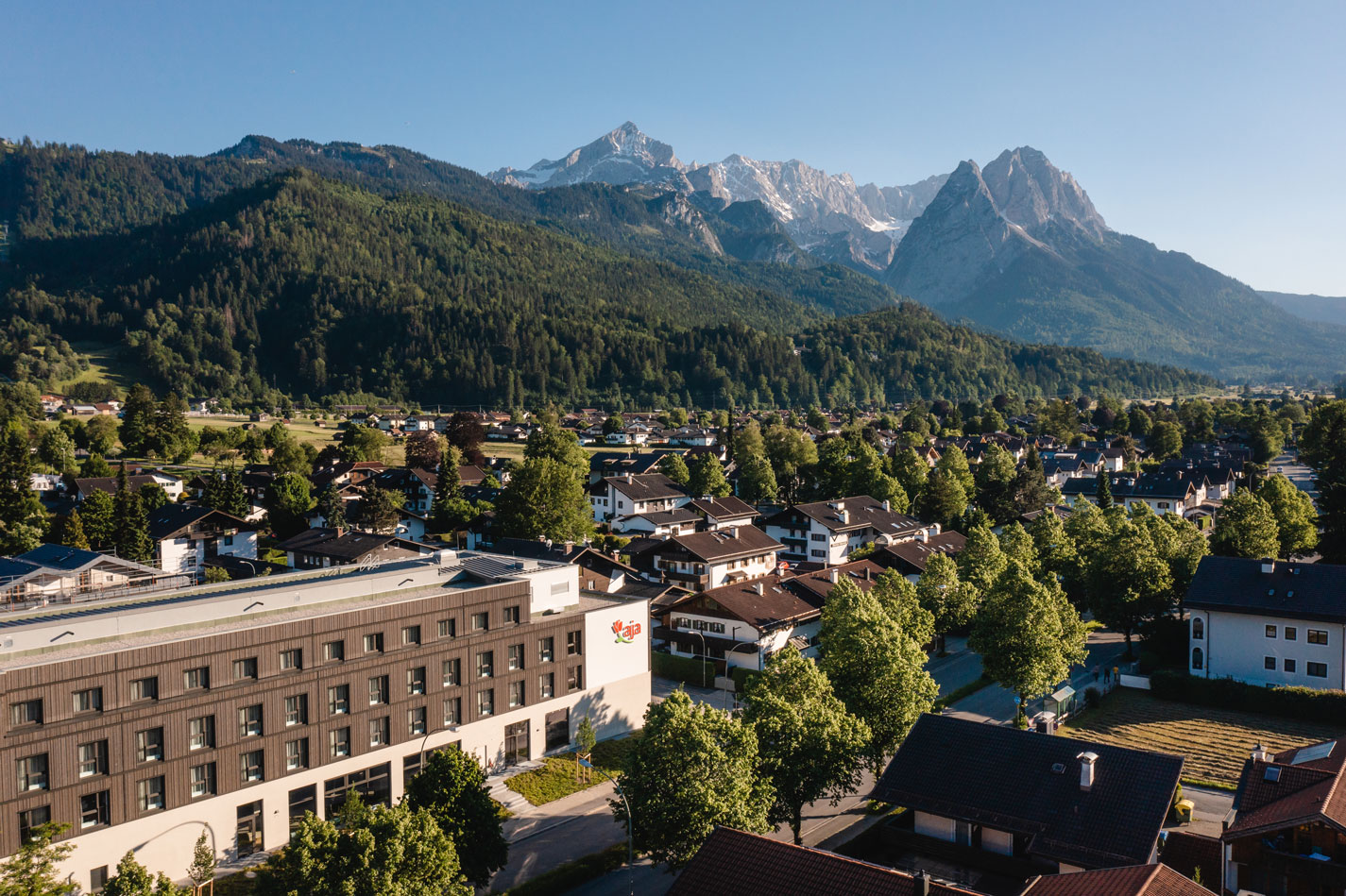 Blick von oben auf das aja Hotel Garmisch-Partenkirche, im Hintergrund bewaldete Hügel und Berggipfel