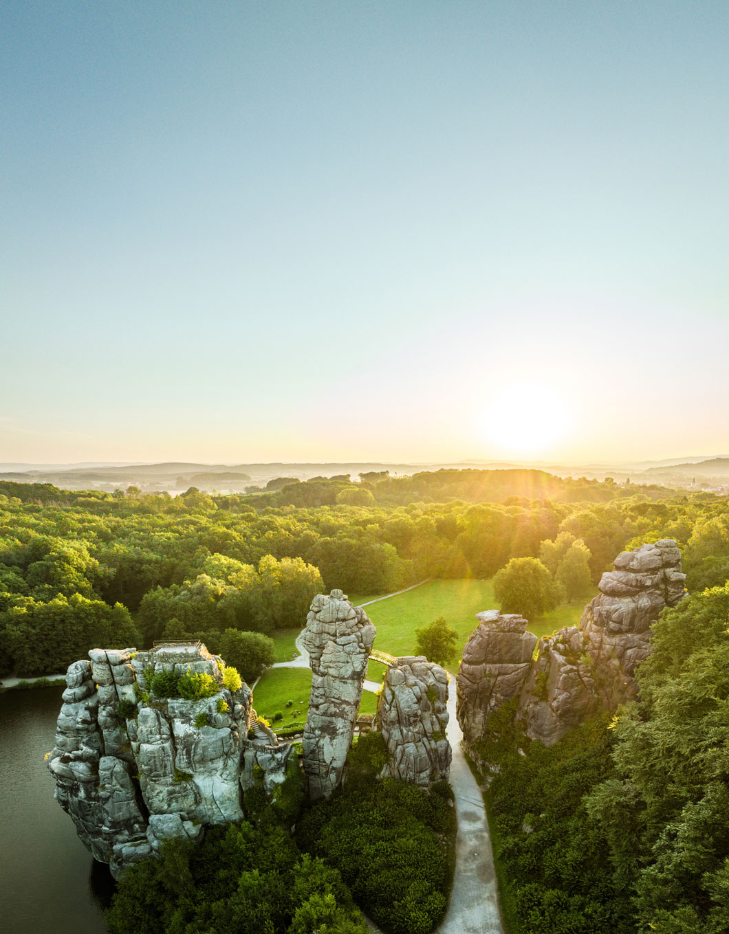 Blick von oben auf markante Felstürme im Sonnenlicht, dahinter erstrecken sich eine grüne Waldlandschaft und der Himmel.