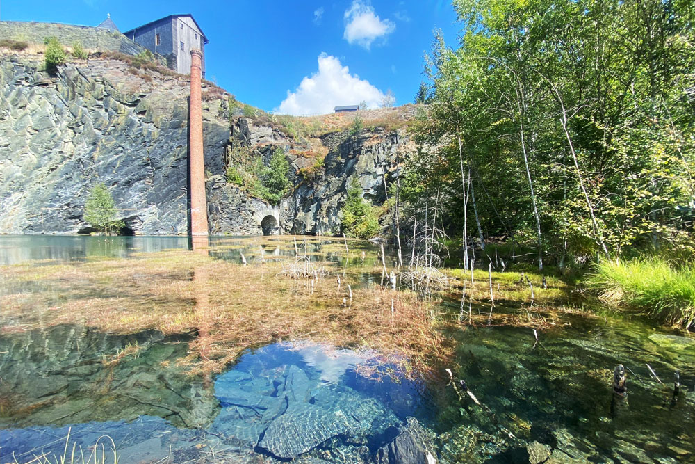 Klares Gewässer, in dem Wasserpflanzen wachsen und Steine zu sehen sind,mit Felswand und Industrieturm im Hintergrund