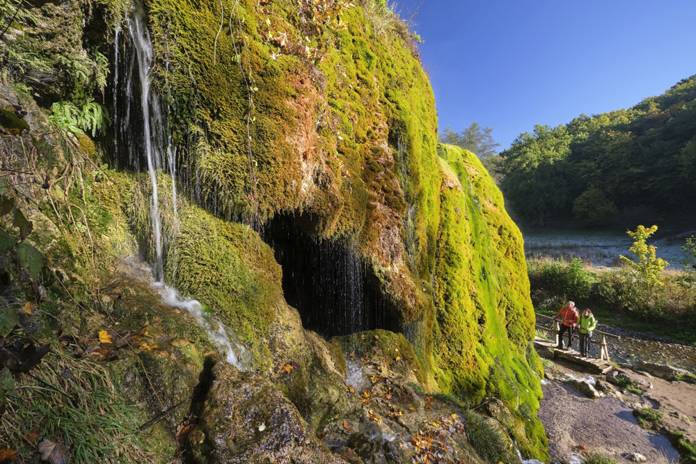 Moosbewachsener Fels an dem ein Wasserrinnsaal herunterfließt, unterhalb blicken zwei Personen von einem Weg zum Wasserfall empor.