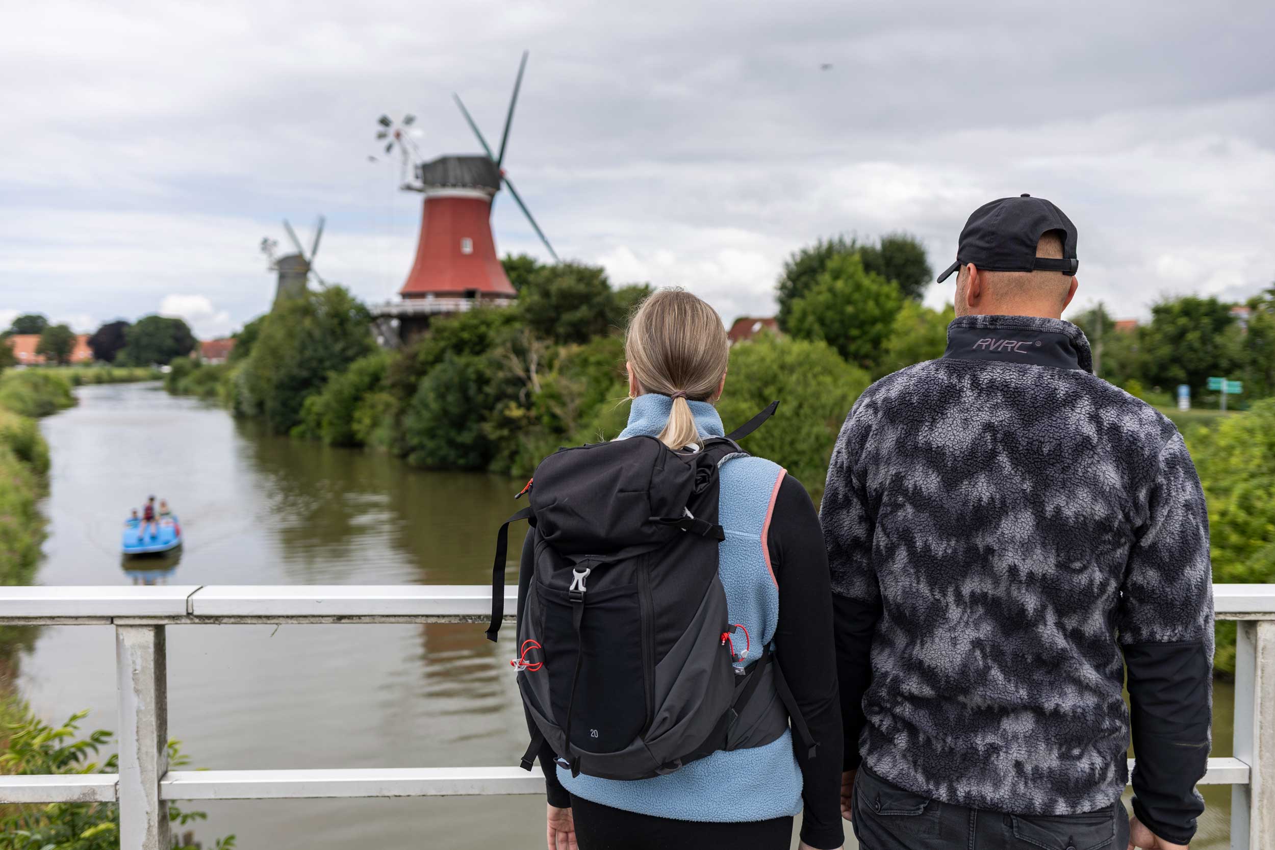 2 Wandernde stehen auf einer Brücken und schauen auf einen Fluss und eine Windmühle