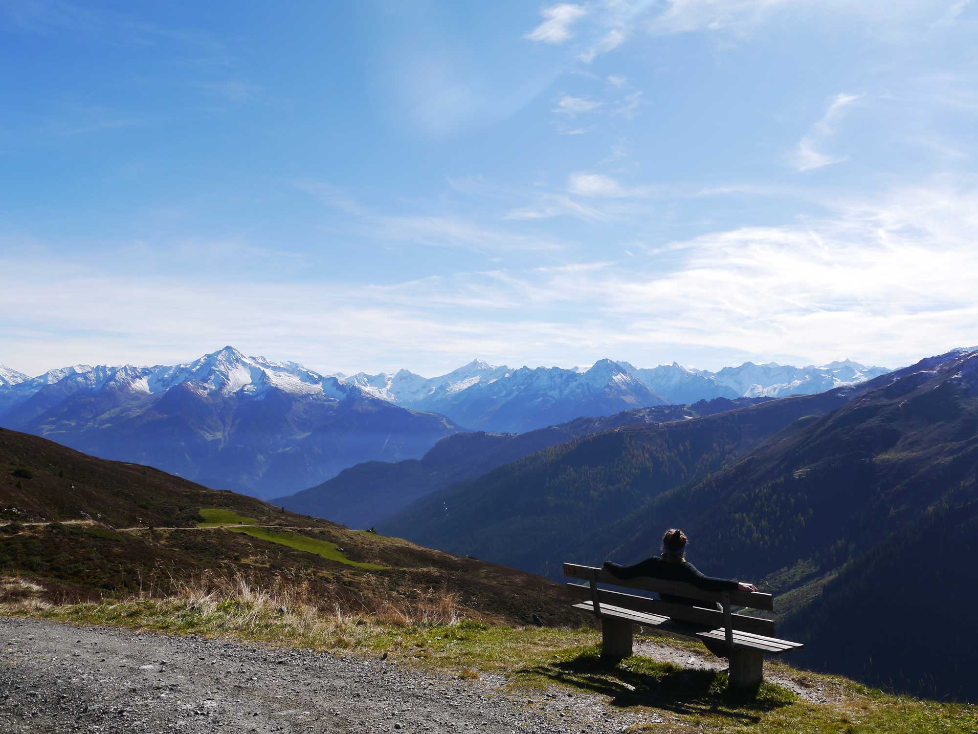 Eine Person sitzt auf einer Bank am Wanderweg und genießt den Ausblick auf die Alpen