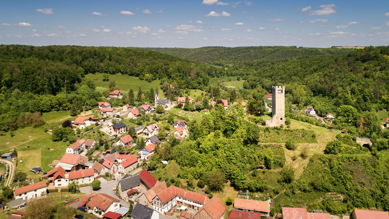 Blick von oben auf die Häuser einer Ortschaft, rote Dächer und Turm, rundherum bewaldete Hügel