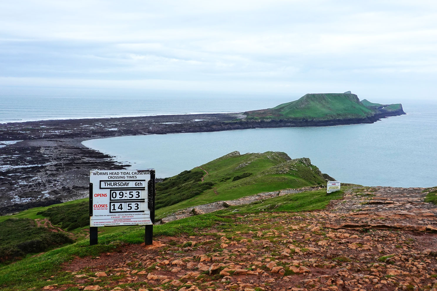 Blick auf eine dünne Landspitze im Meer und eine Hinweistafel zu den Gezeiten