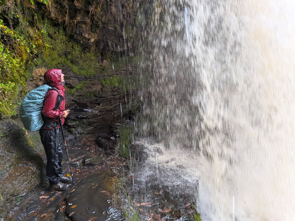 Nasse Wanderin in Regenkleidung neben einem Wasserfall