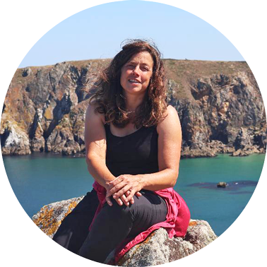 Eine Frau mit windzerzaustem Haar sitzt auf einem Stein an der Küste