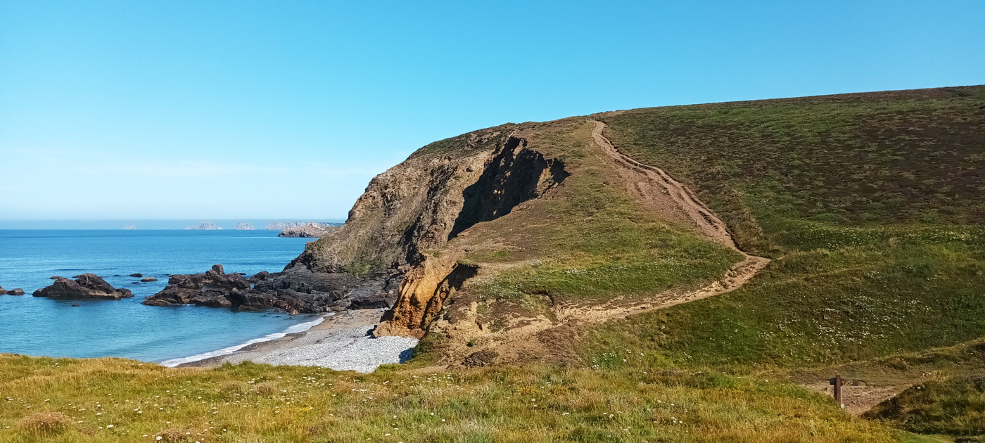 Ein schmaler Pfad auf einer mit Gras bewachsenen Klippe und eine kleine steinige Bucht.