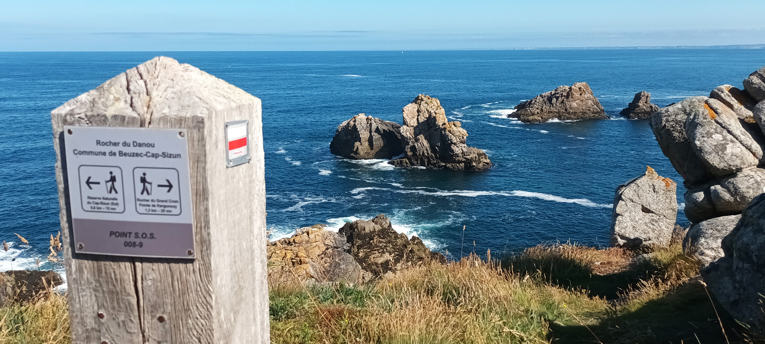 Ein Pfahl mit Wanderwegmarkierungen steht vor einer Meeresbucht mit schroffen Felsen im Wasser