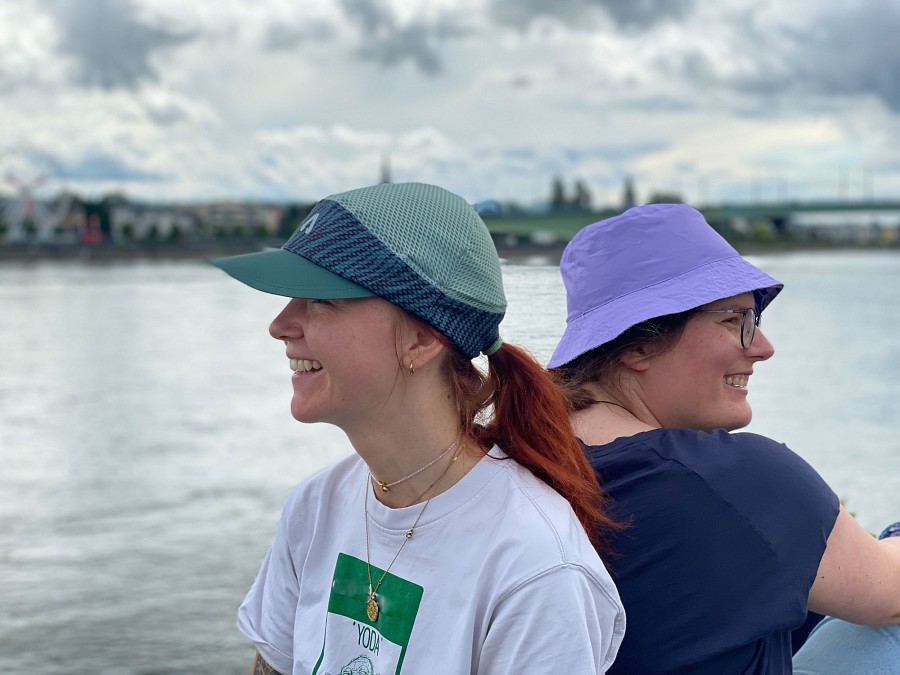 Zwei Frauen sitzen nebeneinander und tragen eine grüne Cap und einen violetten Sonnenhut