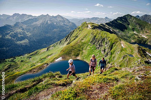 Blick auf einen Bergsee, grüne Berghänge und zwei Wandernde