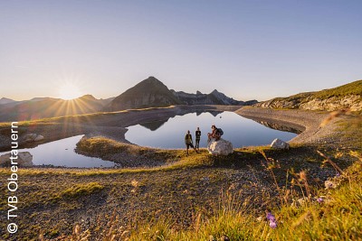 Blick auf spitze Gipfel und einen Bergsee mit Wandernden im Abendlicht