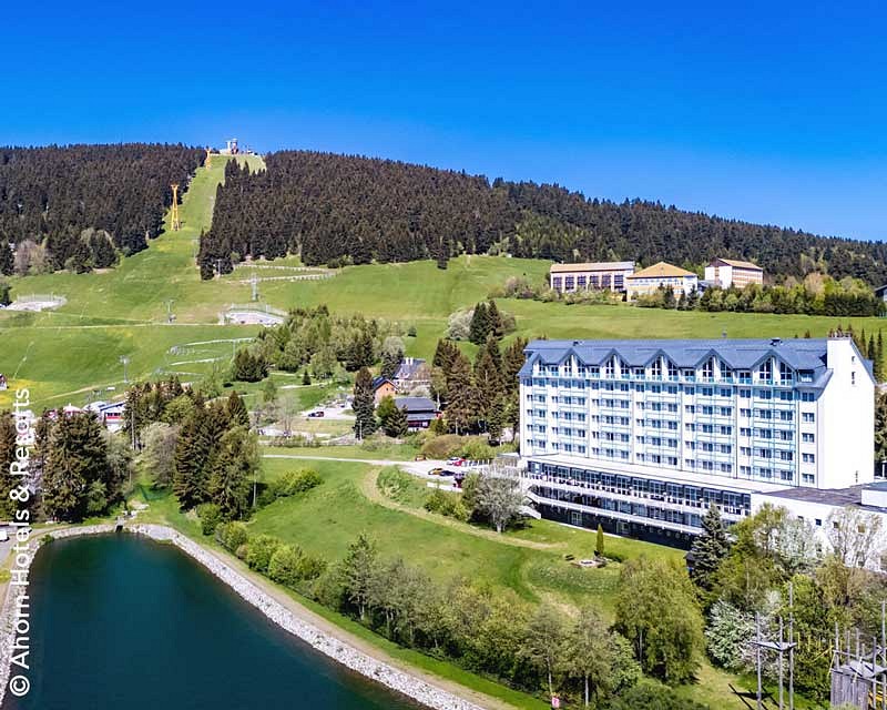 Das Hotel Ahorn Hotel Oberwiesenthal vor der Kulisse des Fichtelbergs und neben einem Stausee. 