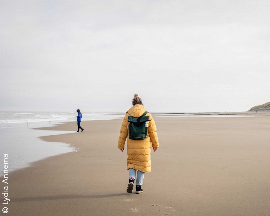 Eine Frau spaziert in dicker Jacke am Strand entlang. 