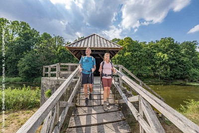 Ein Wanderer und eine Wanderin gehen über eine Holzbrücke auf die Kamera zu, neben ihnen ist ein Gewässer