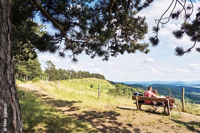 Neben einem Wanderweg sitzt ein Paar auf einer Bank am Rand einer Klippe und schaut in die Ferne auf grüne Hügel