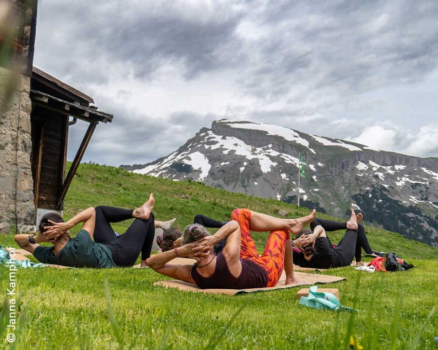 Menschen bei einer Yogaübung auf grüner Wiese, im Hintergrund Bergkulisse