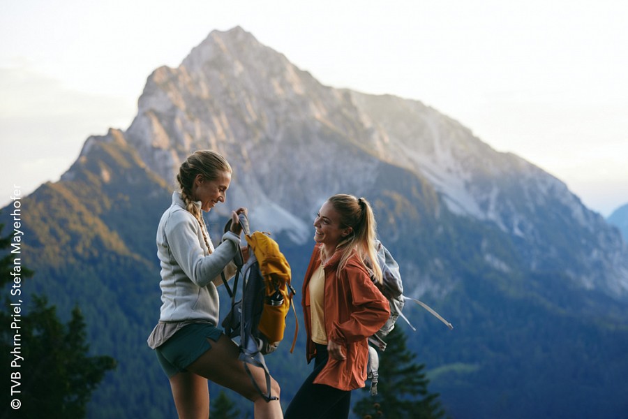 Zwei Freundinnen schultern ihre Rucksäcke vor einem imposanten Berggipfel.