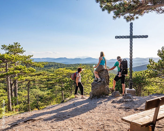 Personen an einem Gipfelkreuz blicken auf den Wald und die Bergkette vor ihnen
