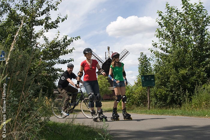 Eine Frau und ein Mädchen fahren auf Inline-Skates, ein Mann fährt auf einem Fahrrad, im Hintergrund befindet sich eine Windmühle. 