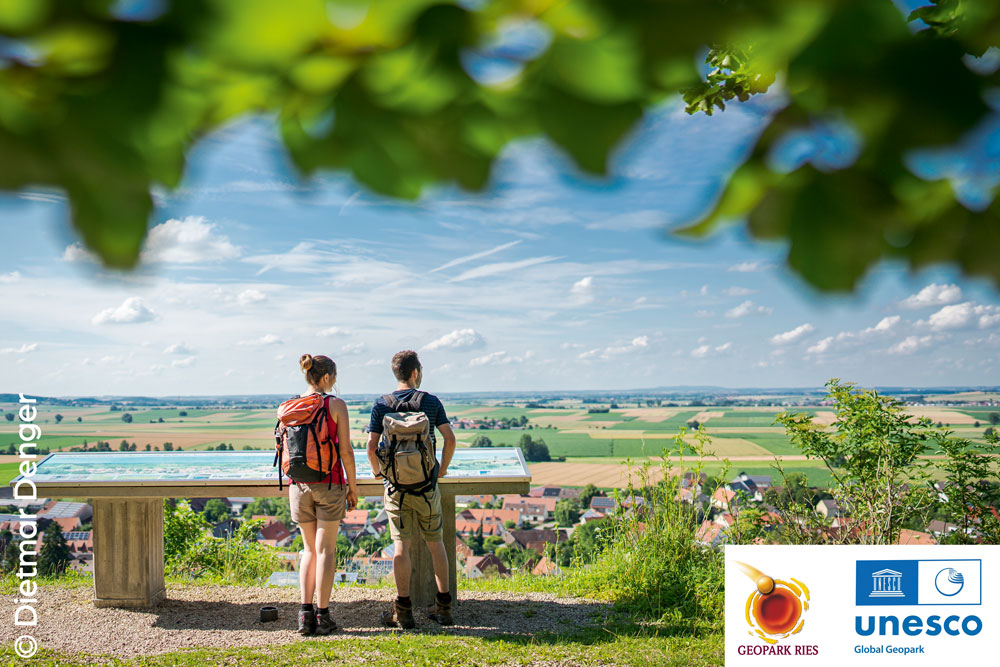 Zwei Wandernde an einer Infotafel blicken über eine weite Feld- und Wiesenlandschaft