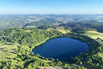 Ein rundes blaues Gewässer von oben, eingebettet in dicht bewaldeten Landschaft.
