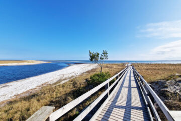 Ein Holzsteg führt am Wasser entlang durch Gräser bis zum Strand