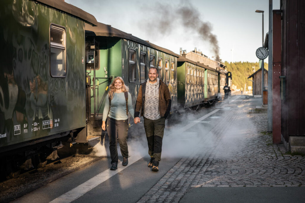 Zwei Wandernde neben historischen Eisenbahnwagons, im Hintergrund steigt Dampf aus der Lokomotive auf.