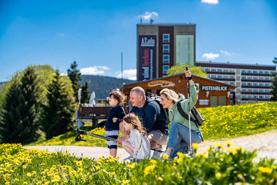 Eine Familie hockt am Rande einer Blumenwiese und blickt in die Ferne, im Hintergrund ein Hotel vor blauem Himmel.