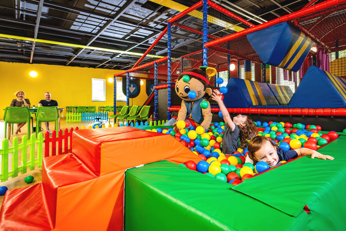 Spielende Kinder und ein Maskottchen in einem Bällebad umringt vom bunten Polstern. 