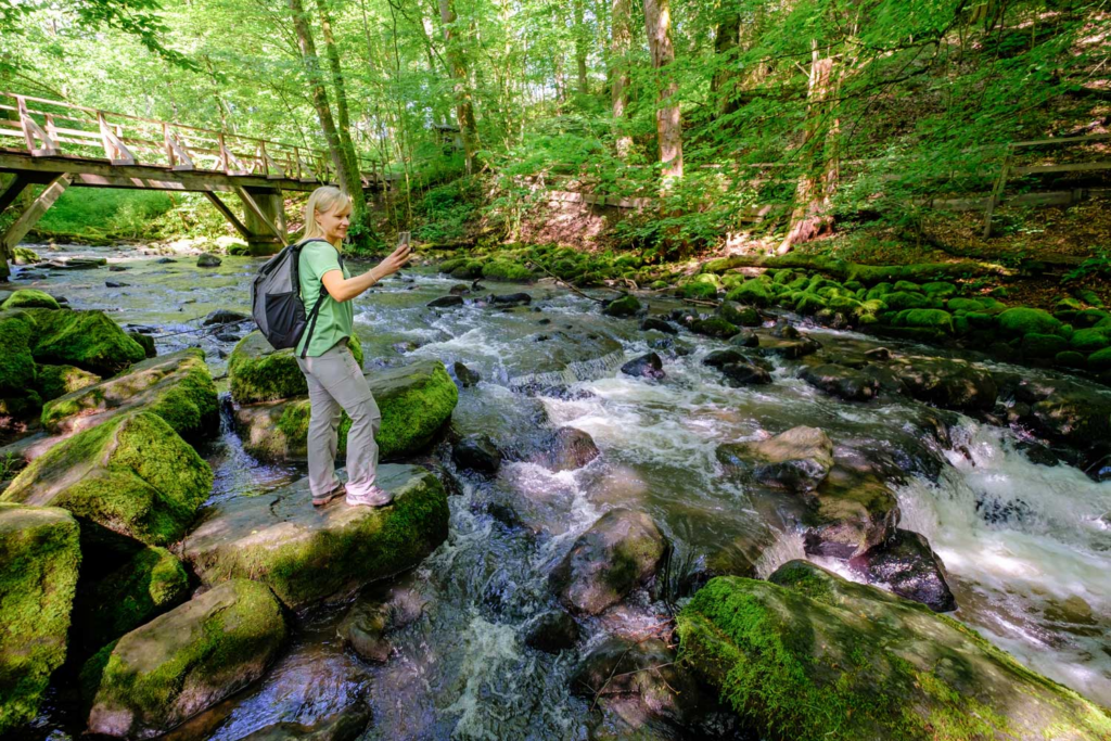 Eine Frau steht fotografierend auf einem Stein am Flussufer, sie ist umgeben von Wald, im Hintergrund eine Holzbrücke.
