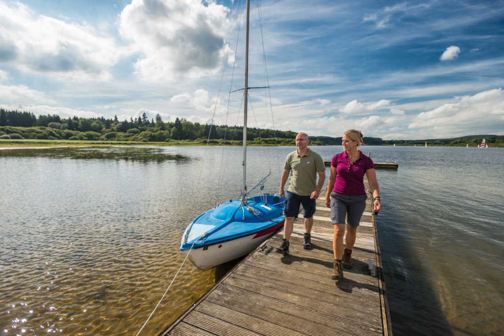 Zwei Wandernde kommen über einen Steg auf die Kamera zu, umgeben von Wasser und einem festgemachten Boot.