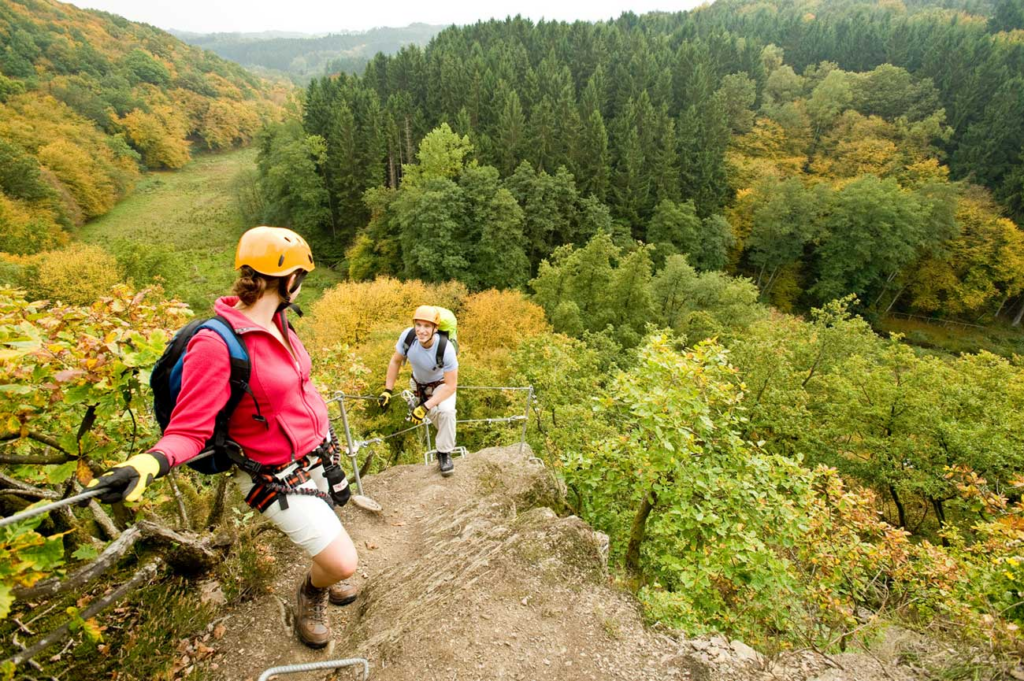 Zwei Wandernde mit Klettersteigset und Helm an einem steinigen und schmalen Pfad, unter ihnen einen leicht herbstlich gefärbte Waldkulisse