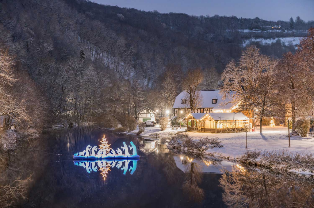 Ein weihnachtlich beleuchtetes Ensemble auf einem Fluss in der Dämmerung, daneben ein mit Lichterkette geschmücktes Fachwerkhaus
