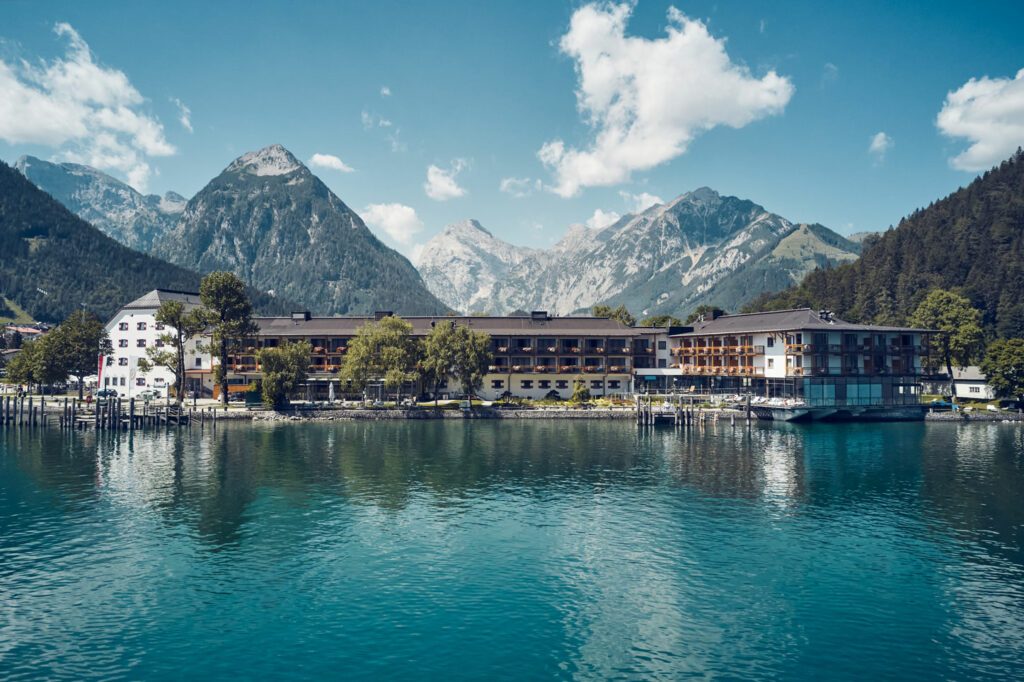 Leucht blaues Wasser, an dessen Ufer ein Hotelgebäude und Berggipfel dahinter.