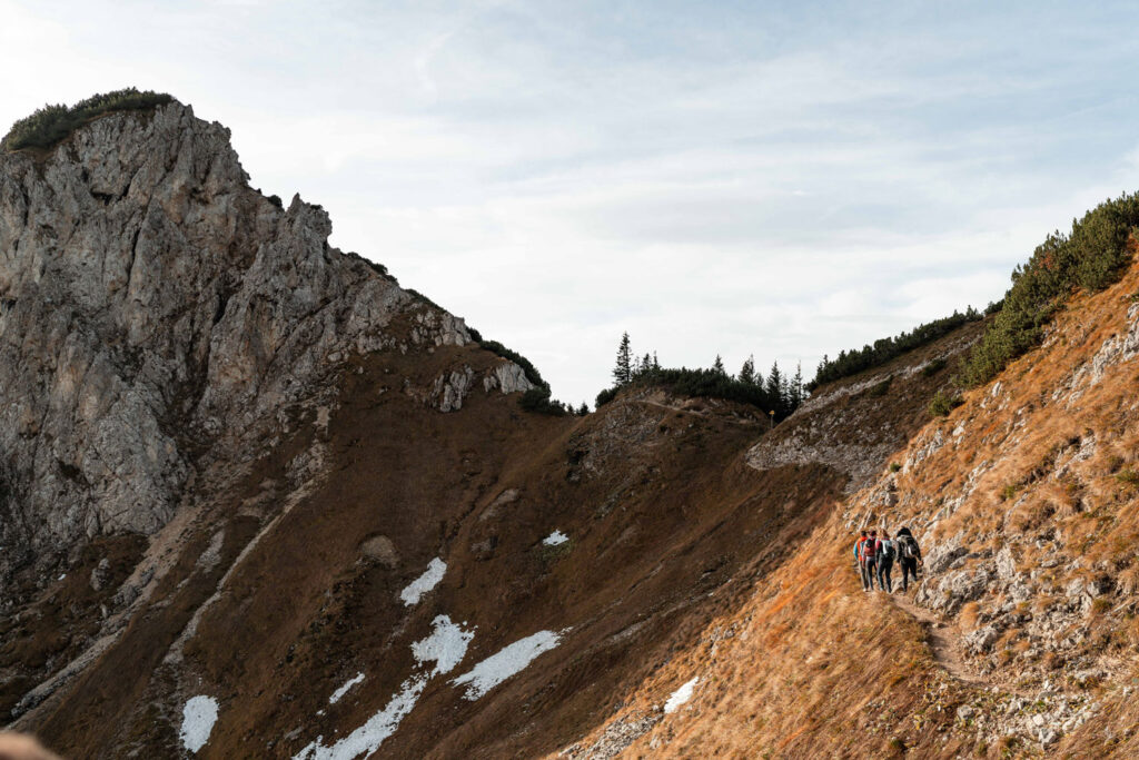 Eine Felsenwand und kleine Schneefelder. Daneben ein schmaler Pfad auf dem eine Wandergruppe entlangläuft