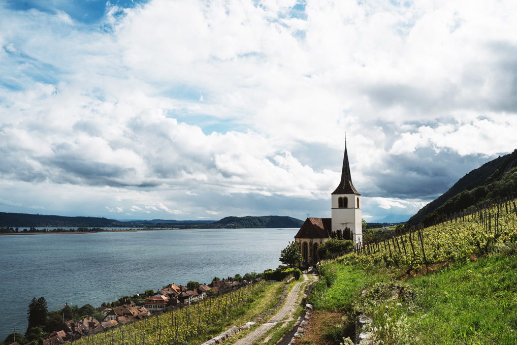 Eine weiße Kirche steht zwischen Weinberg und Dorf, im Hintergrund ein See mit dramatischen Wolken
