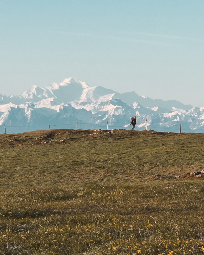 Ein Wanderer in der Ferne auf einer grünen Anhöhe, im Hintergrund ein schneebedecktes Bergmassiv