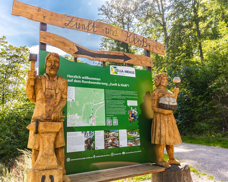 Ein großes Schild und zwei große Holzfiguren mit Infos und dem Namen des Wanderwegs