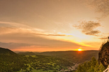 Eine Person steht auf einem Felsvorsprung, vor ihr ein tiefer und weiter Ausblick über eine Hügellandschaft im Sonnenuntergang