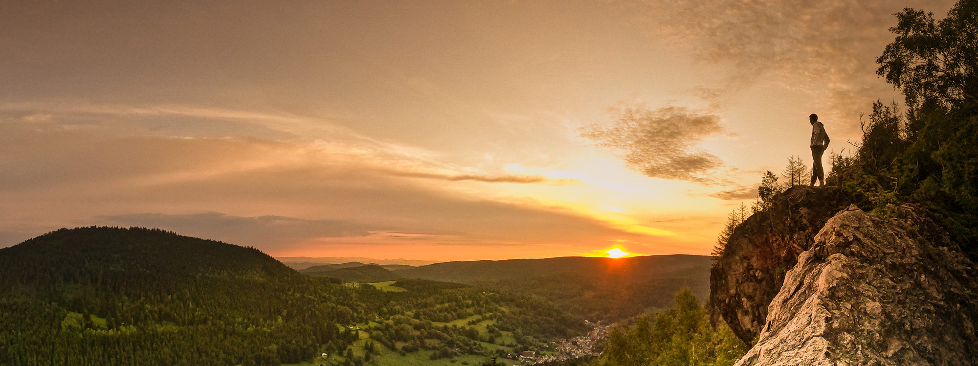 Eine Person steht auf einem Felsvorsprung, vor ihr ein tiefer und weiter Ausblick über eine Hügellandschaft im Sonnenuntergang