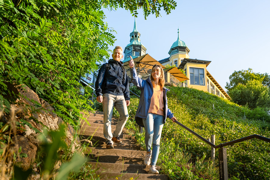 Zwei Wandernde im Dresden Elbland kommen auf Treppen einen grünen Hang hinab, im Hintergrund ein Gebäude mit hellblauen spitzen Türmen.